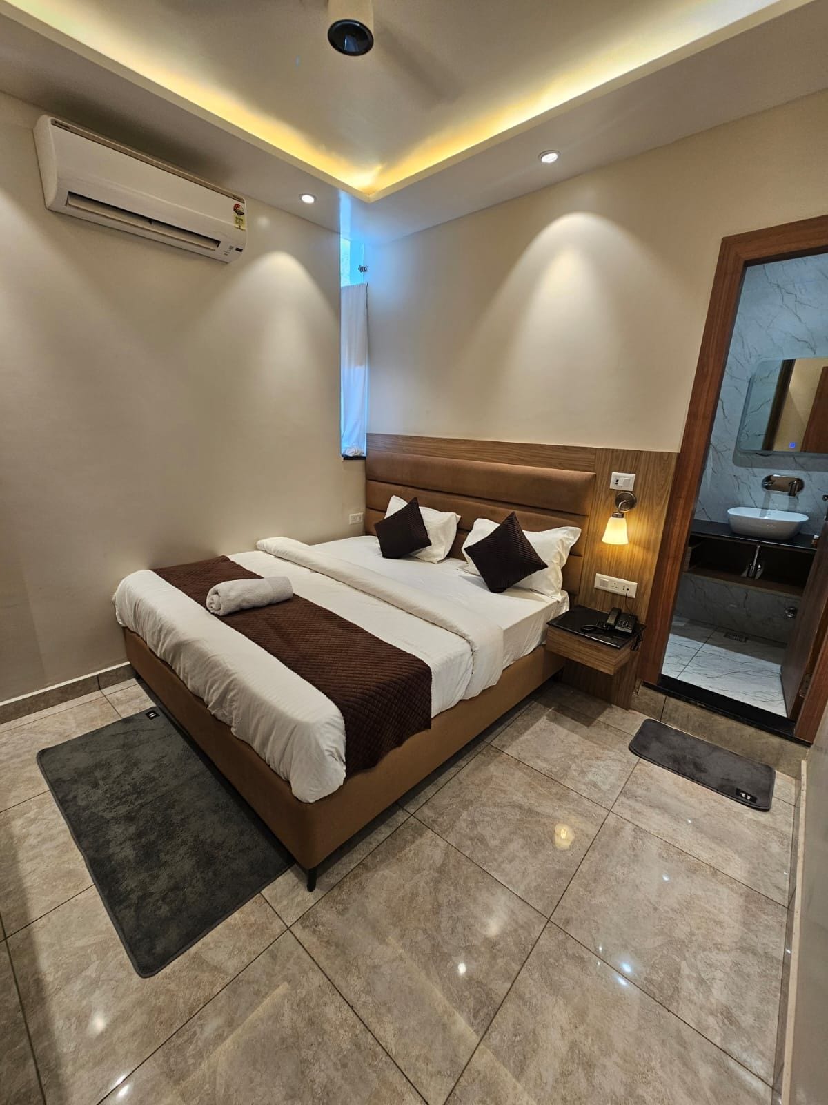 A hotel room with a bed, bedside table, lamp, air conditioning unit, and a bathroom mirror.