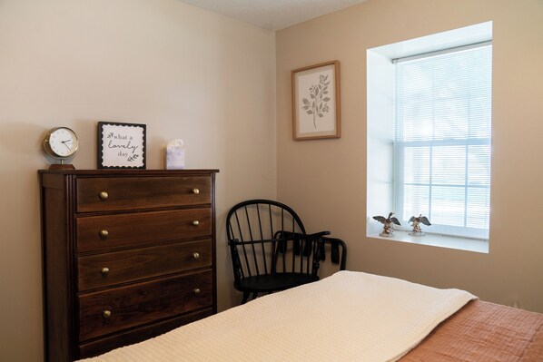 A bedroom with a wooden dresser, a chair, a bed with a white blanket, a wall clock, and a window with blinds.