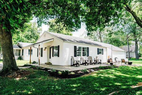 A white single-story house with a porch, a covered outdoor seating area with Adirondack chairs, and a well-maintained lawn.