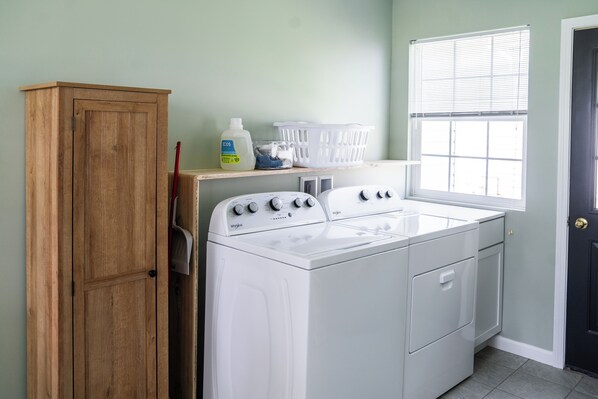 A laundry room with a white washer and dryer, wooden cabinet, laundry baskets, and a window with blinds.