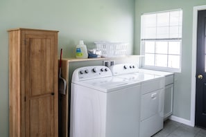 A laundry room with a white washer and dryer, wooden cabinet, laundry baskets, and a window with blinds.