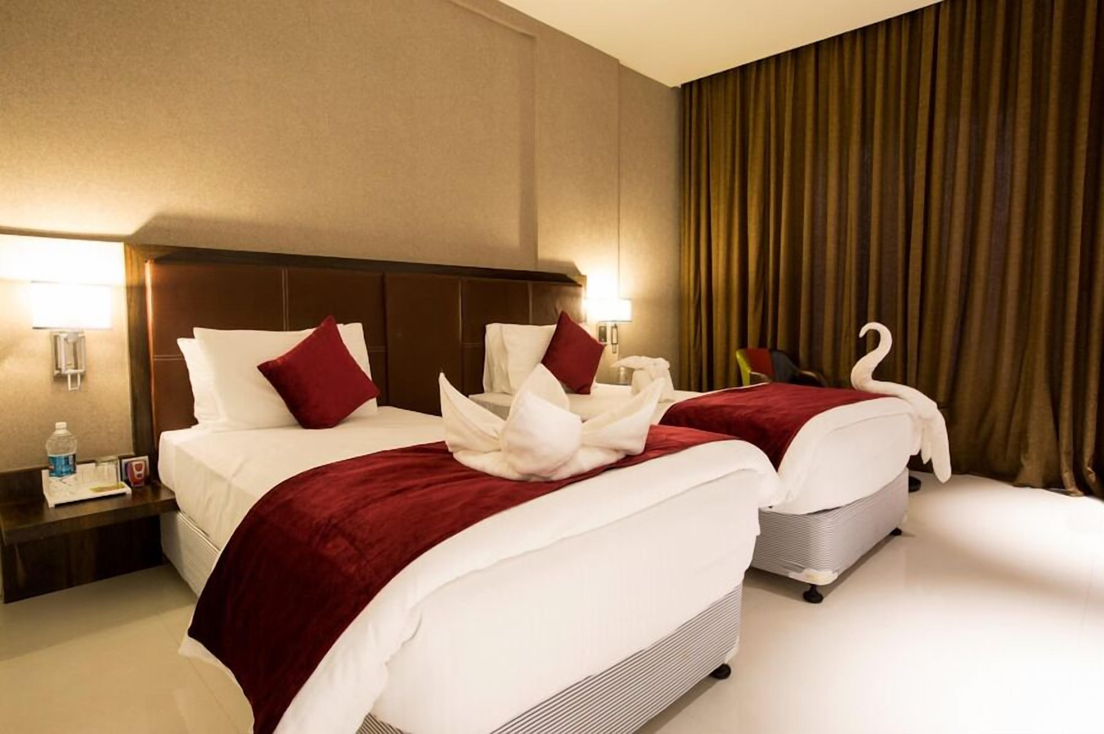 Habitación de hotel con dos camas, cada una con una colcha roja y almohadas blancas, un cabecero de madera y una mesita de noche con una lámpara y una botella.