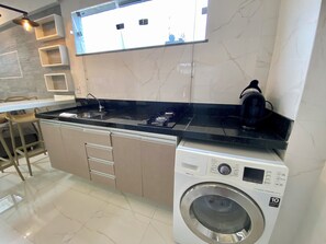 A compact kitchen with a built-in washing machine, a bar stool, and a hanging light fixture. - 307 2Qtos Funcional bem Localizado (Belém)