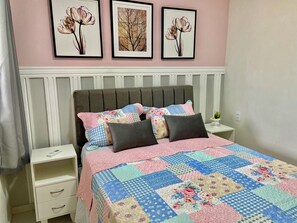 Un dormitorio con cama, almohadas, una mesita de noche y una pared decorada con cuadros.
