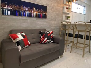 Un salón moderno con un sofá gris, dos almohadones estampados, un cuadro de un paisaje urbano en la pared, un taburete de madera y un suelo de baldosas blancas.