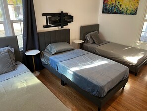 Ein Zimmer mit zwei Einzelbetten, einem kleinen Tisch und einem an der Wand montierten Fernseher.