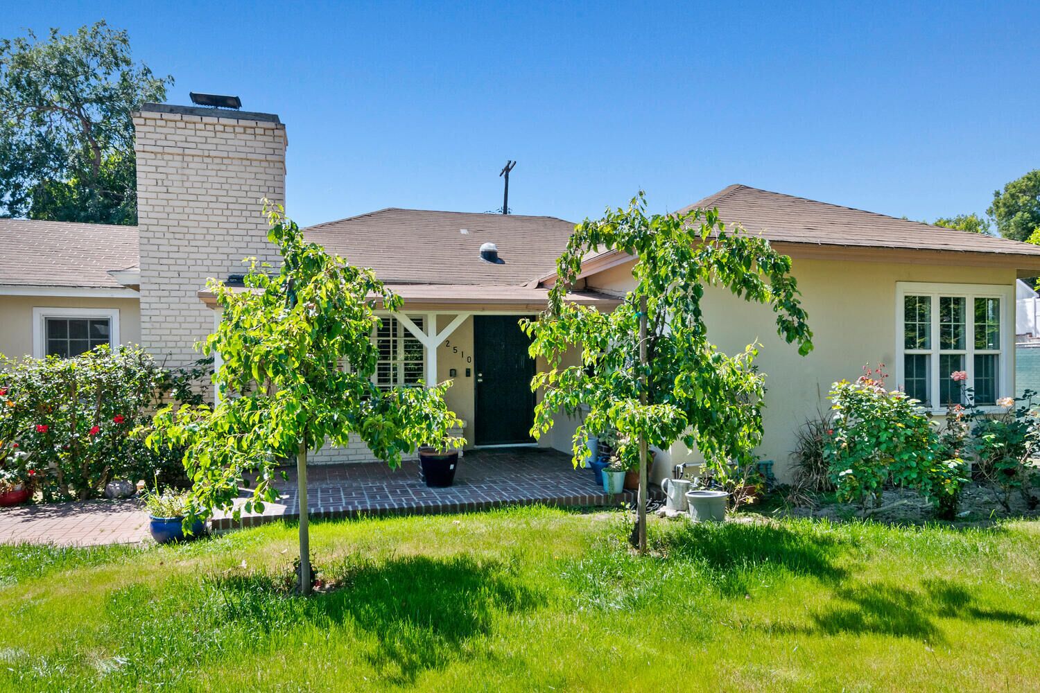 Une maison de plain-pied avec une cheminée en briques, un porche couvert et un jardin bien entretenu où poussent diverses plantes et fleurs.