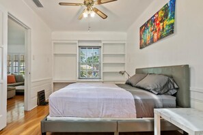 Ein Schlafzimmer mit Bett, Schreibtisch, Deckenventilator, einem Fenster mit Jalousien und einem farbenfrohen Gemälde an der Wand.