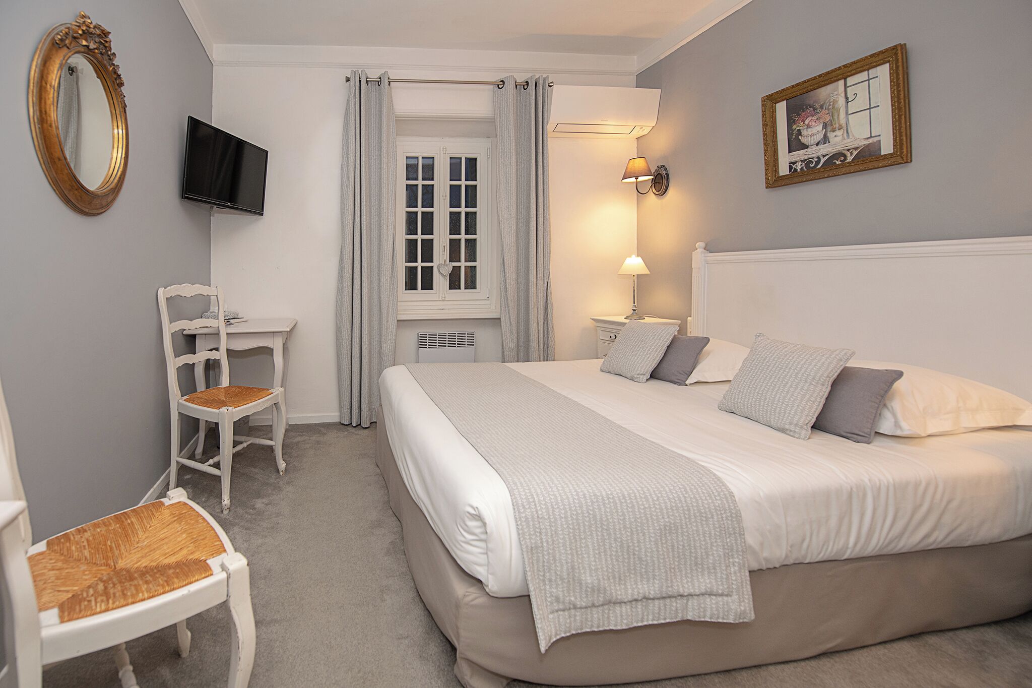 A hotel room with a bed, a chair, a television, a window with curtains, a wall-mounted lamp, and a framed picture.