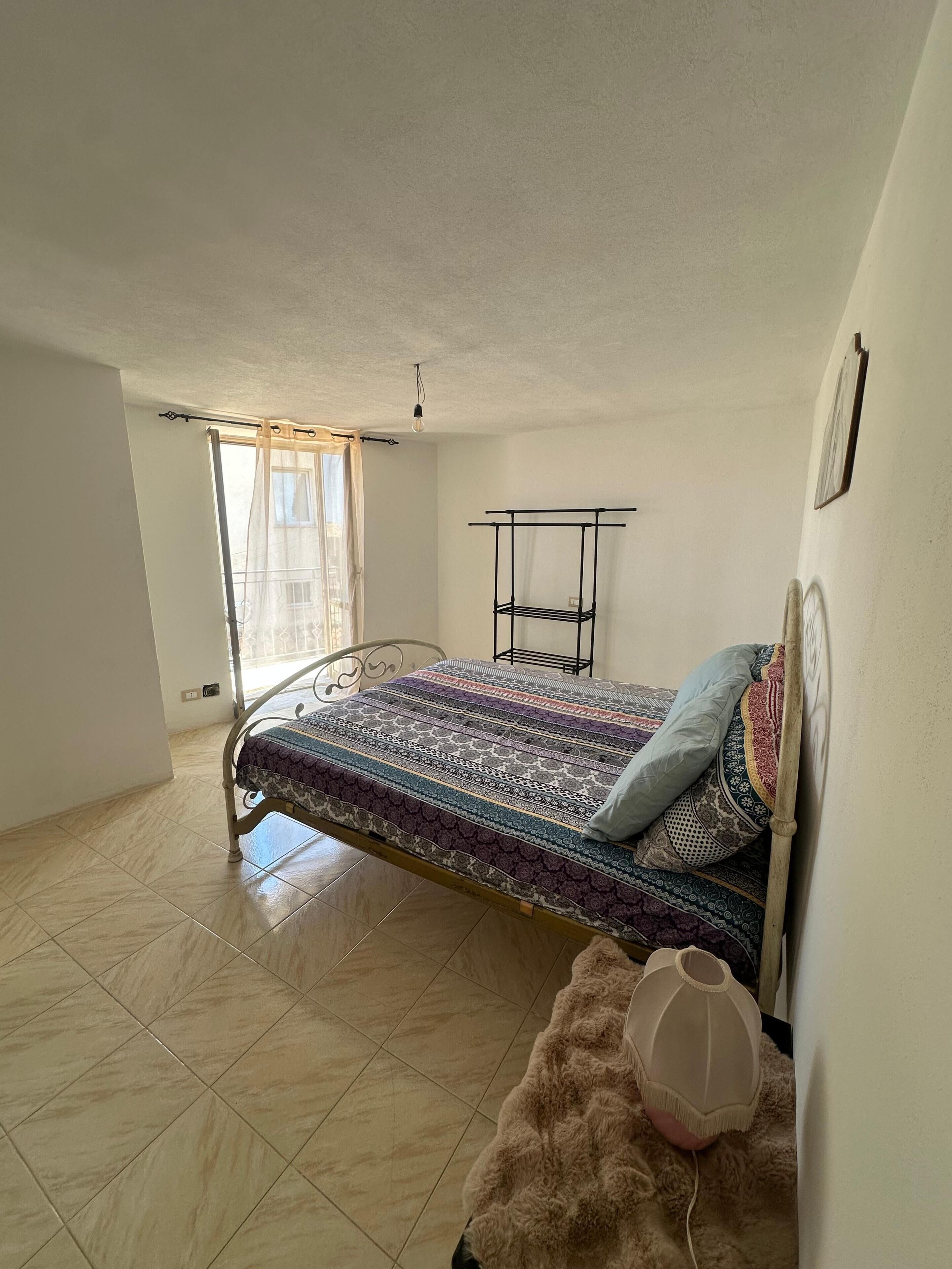 Ein Schlafzimmer mit Bett, einem Fenster mit Vorhängen, einem Heizkörper und einem Wandregal.