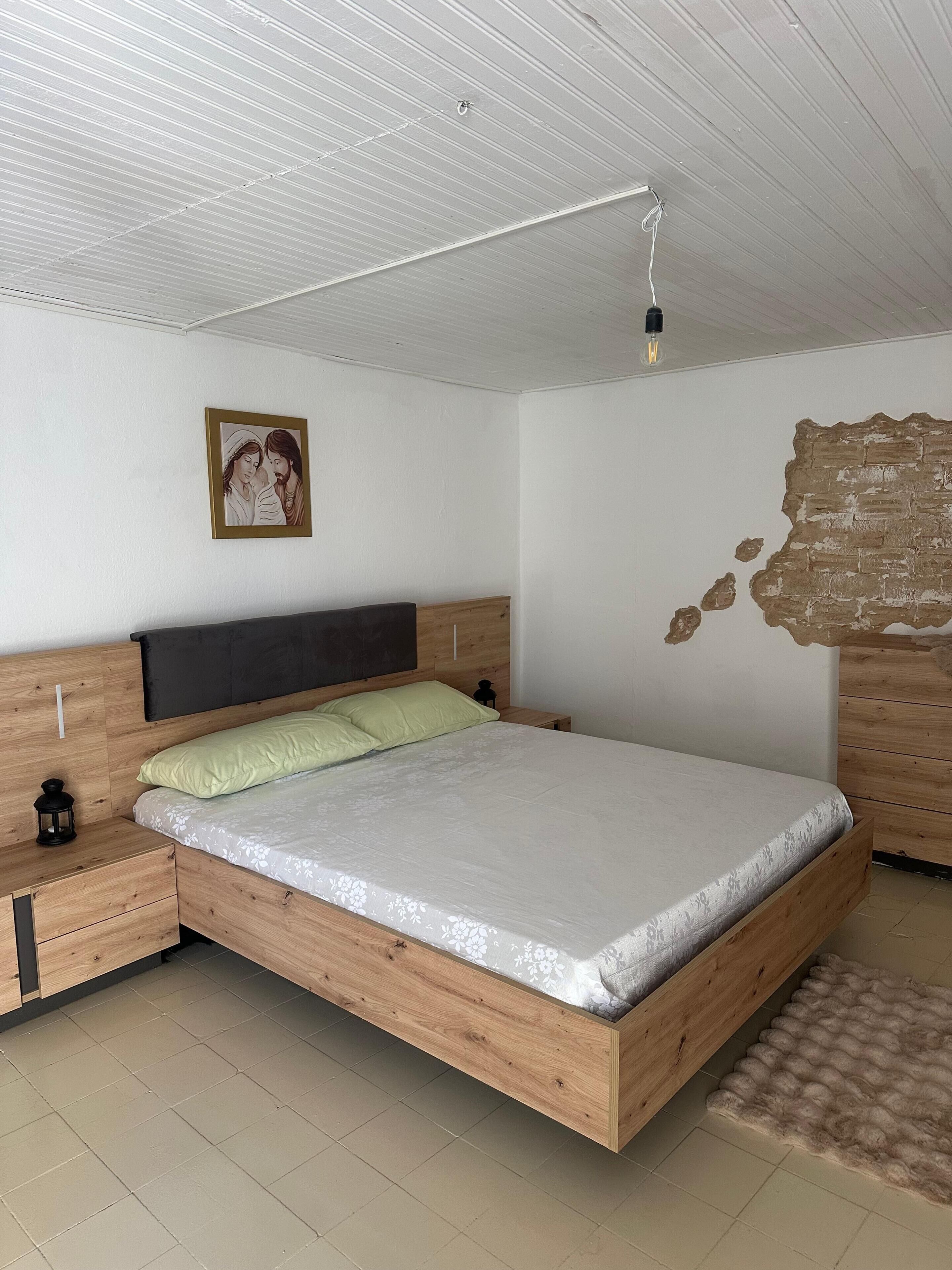 Een slaapkamer met een houten bedframe, een nachtkastje, een wandlamp en een lijst met een afbeelding aan de muur.