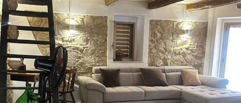 Un salon avec un canapé d’angle, un ventilateur, une lampe et un mur en pierre.