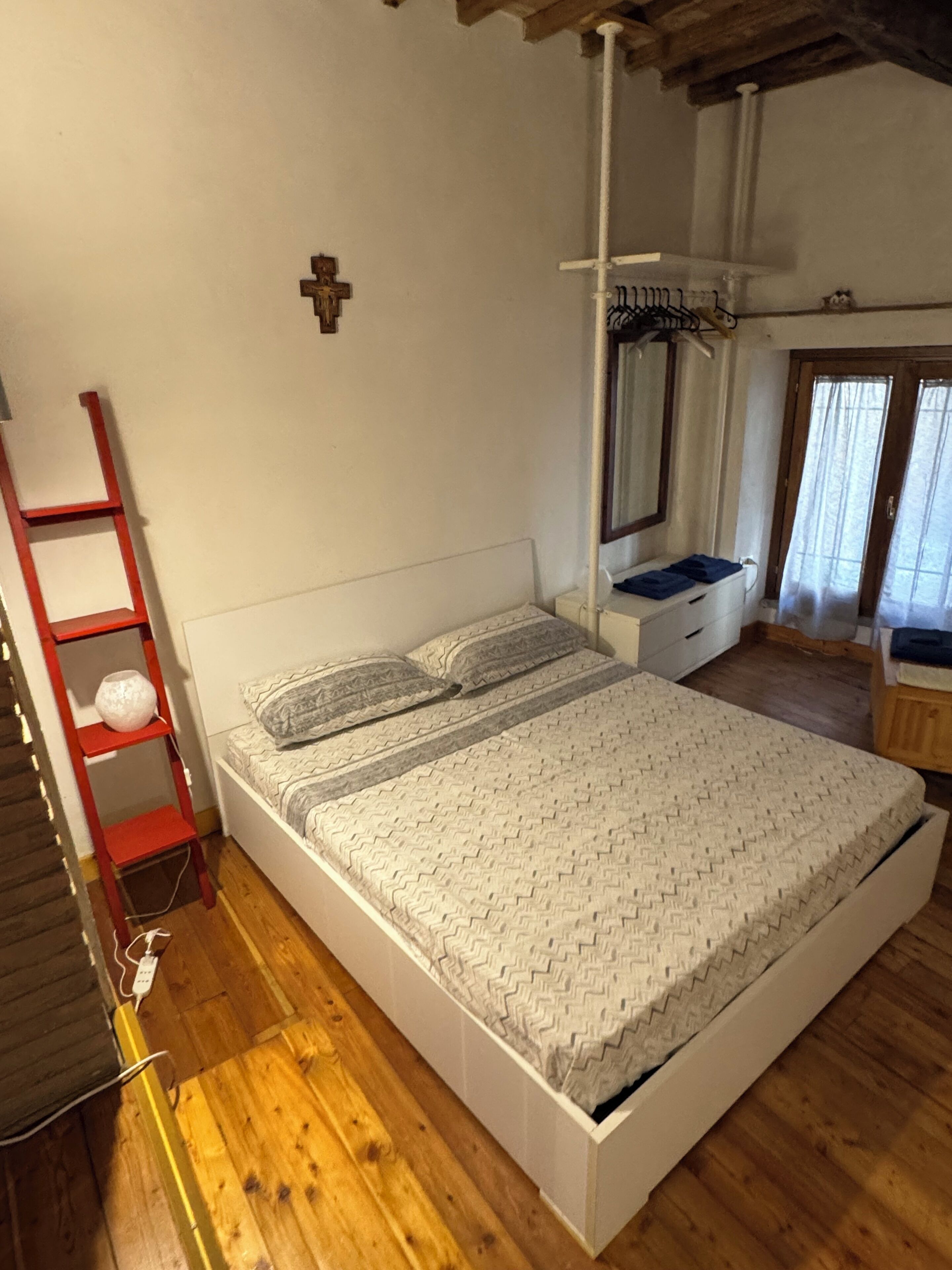 Un dormitorio con piso de madera, cama blanca con ropa de cama estampada, una escalera roja, una repisa montada en la pared y una ventana con cortinas.