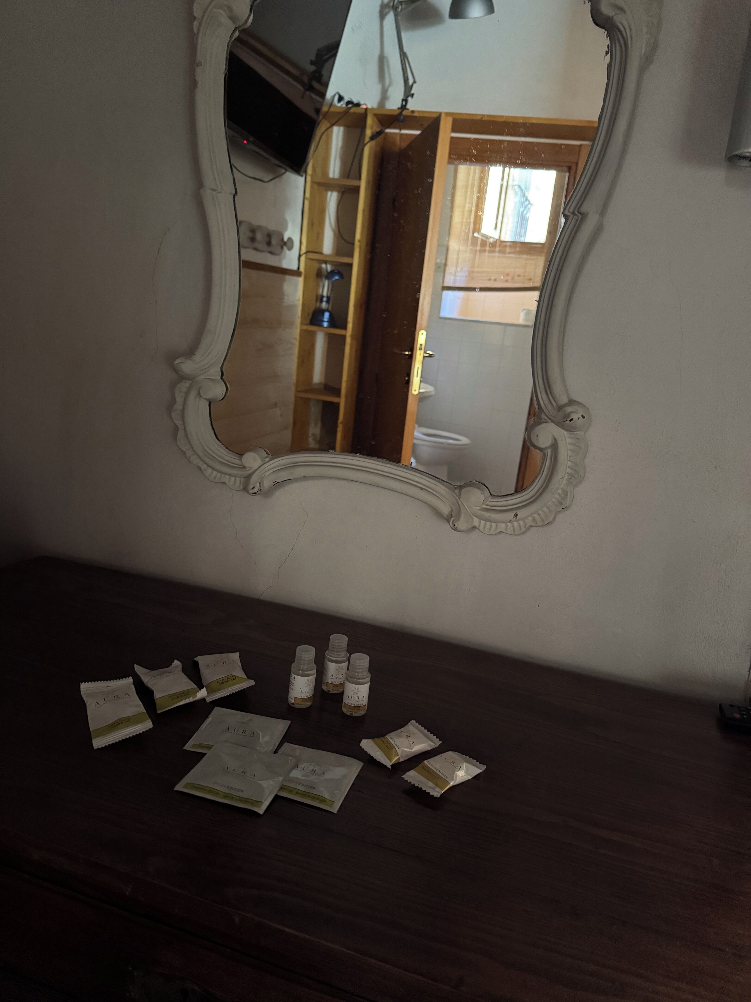 A mirror with a decorative frame on a wall, a wooden dresser below, and a reflection of a room with a door and a window.