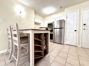 A kitchen with white cabinets, a stainless steel refrigerator, a microwave, and a wooden dining table with chairs. - Natty's Cove Step free/Walk to shops/Heart of Miramar Beach! (Miramar Beach)