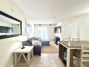 Een gezellige, compacte leefruimte met een bank, een kleine tafel, een aan de muur bevestigde televisie en een eethoek met stoelen.