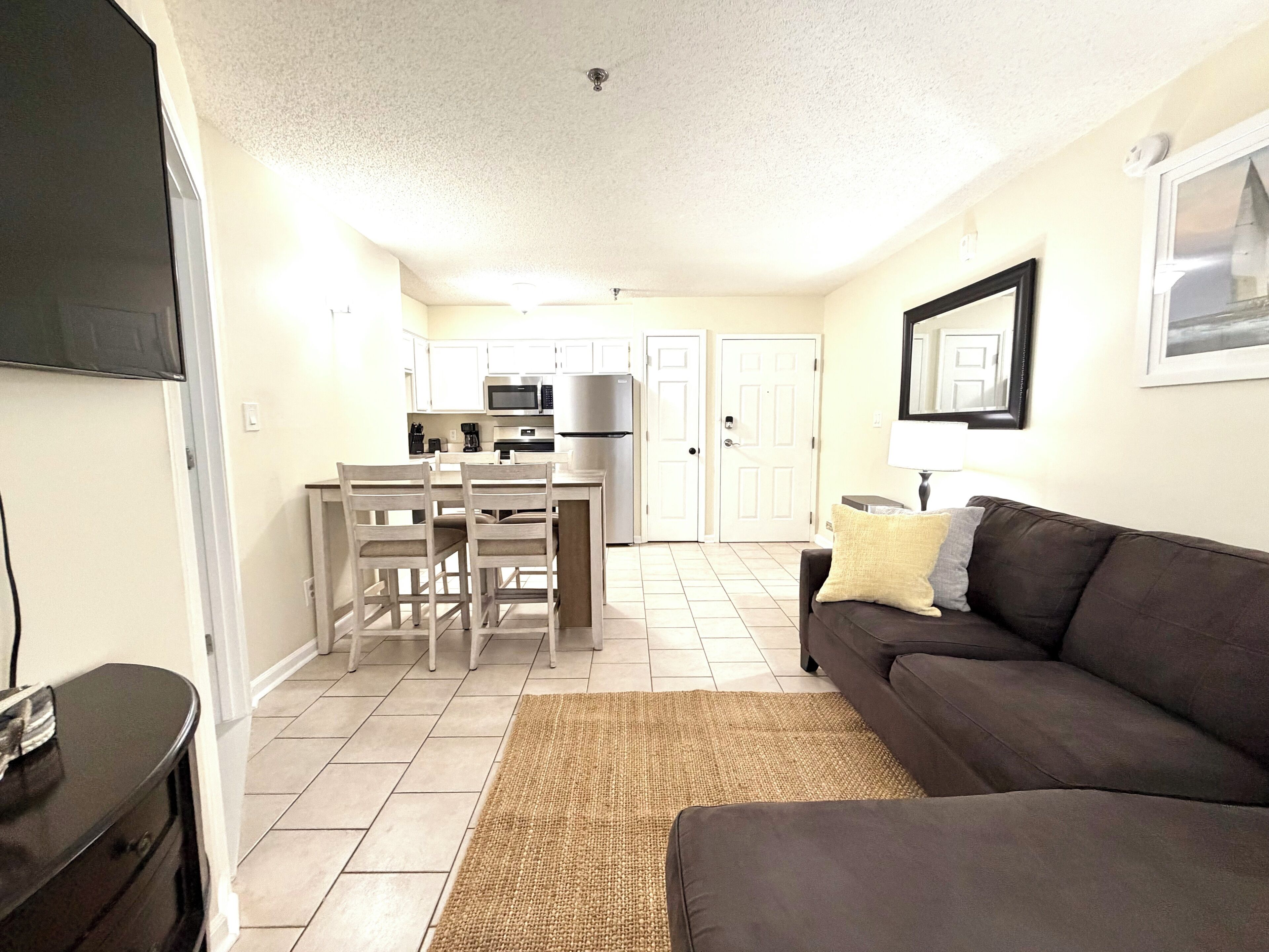 A living room with a dark brown sofa, a coffee table, and a TV on the left. There is a kitchen area with white cabinets, a microwave, and a refrigerator in the background.