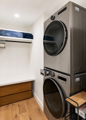 A laundry room with two stacked front-loading washing machines, a wooden bench, and a shelf with folded towels.