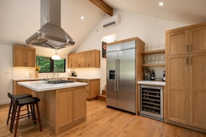 Une cuisine moderne avec des meubles en bois, un réfrigérateur en acier inoxydable, un réfrigérateur à vin et un grand îlot central doté d’une cuisinière à gaz.