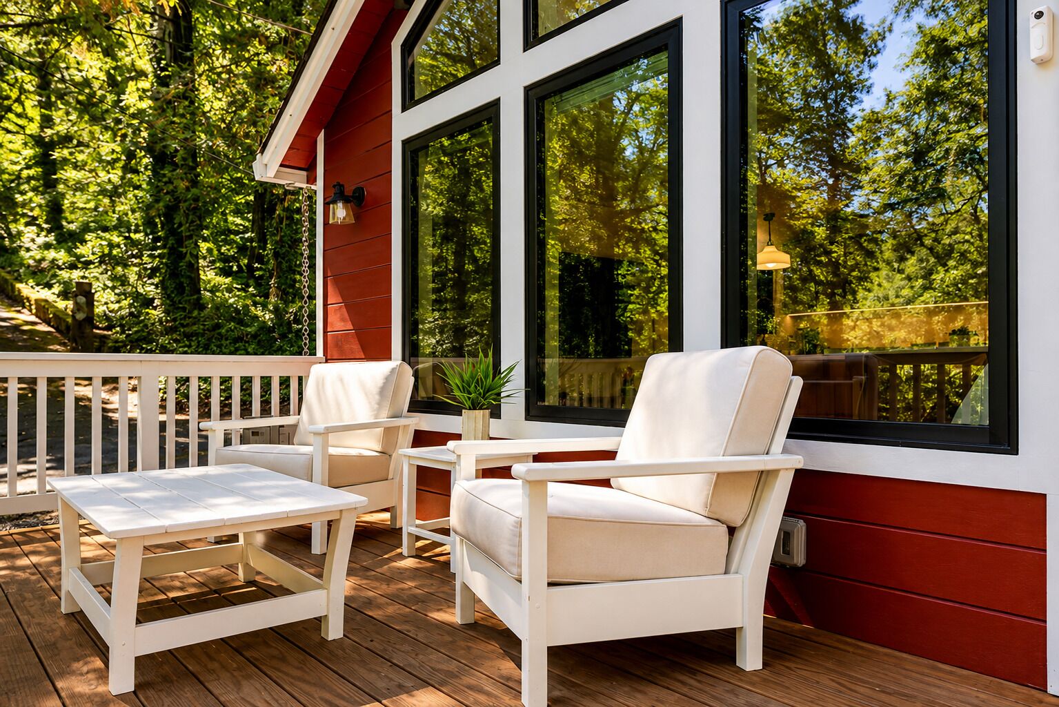 A wooden deck with white outdoor furniture, large windows, and a view of a forested area.