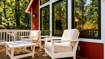 A wooden deck with white outdoor furniture, large windows, and a view of a forested area.