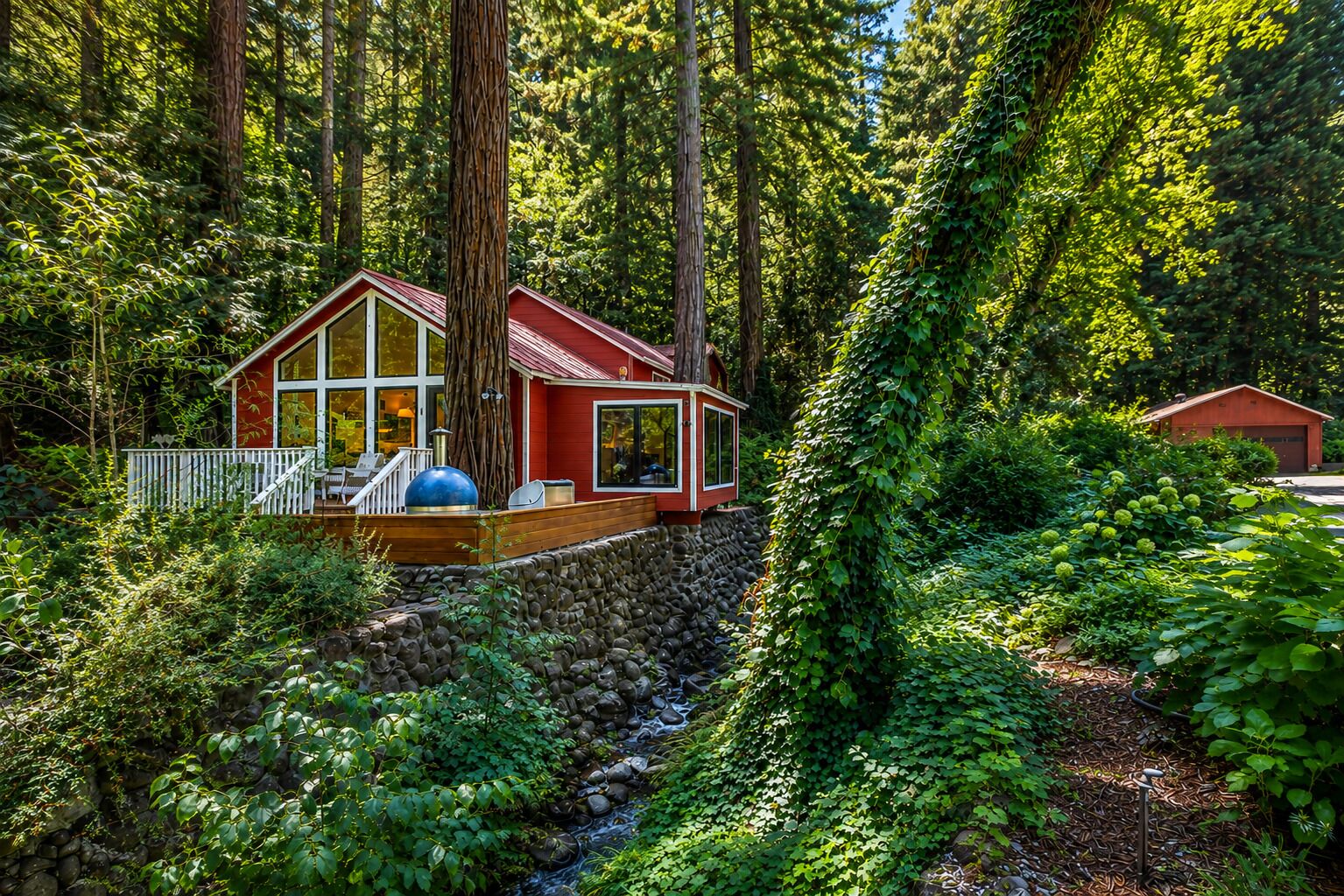 A cabin with a deck, surrounded by dense forest and a stone wall.