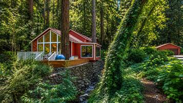 A cabin with a deck, surrounded by dense forest and a stone wall.