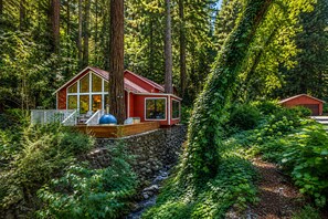A cabin with a deck, surrounded by dense forest and a stone wall. - Storybook Cottage in the Calistoga Redwoods (Calistoga)