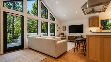 A modern living room with a sofa, a TV, and a kitchen area with wooden cabinets and a large window.
