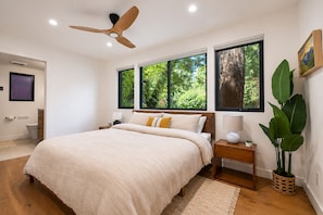 Une chambre avec un lit, un ventilateur de plafond, une fenêtre donnant sur un paysage verdoyant, une table de chevet avec une lampe et une plante en pot.