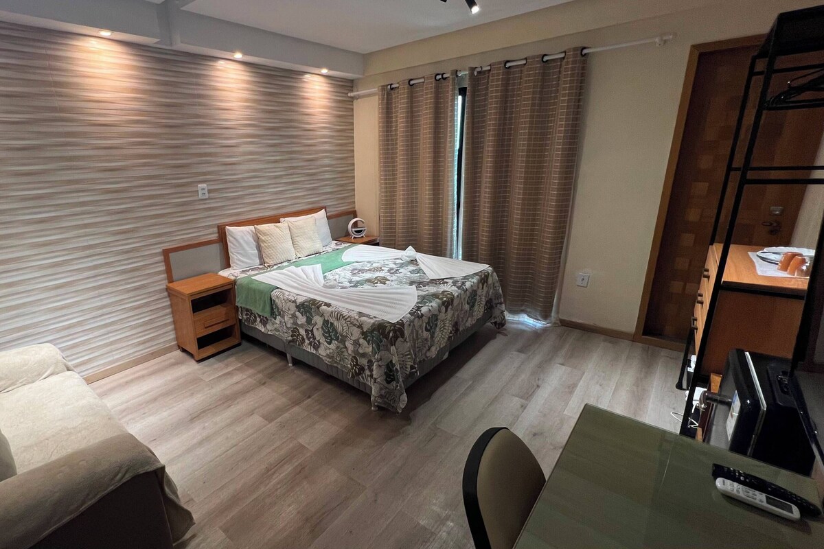 A hotel room with a bed, bedside table, chair, and a desk with a remote control.