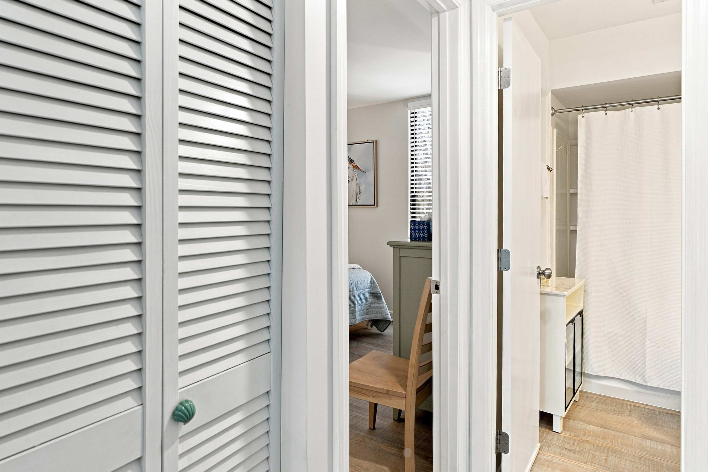 A hallway with a door leading to a bedroom with a bed, a chair, and a window with blinds.