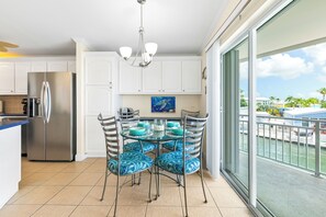 Une cuisine moderne avec des meubles blancs, un réfrigérateur en acier inoxydable et un coin repas comprenant une table en verre et des chaises à motifs bleus.