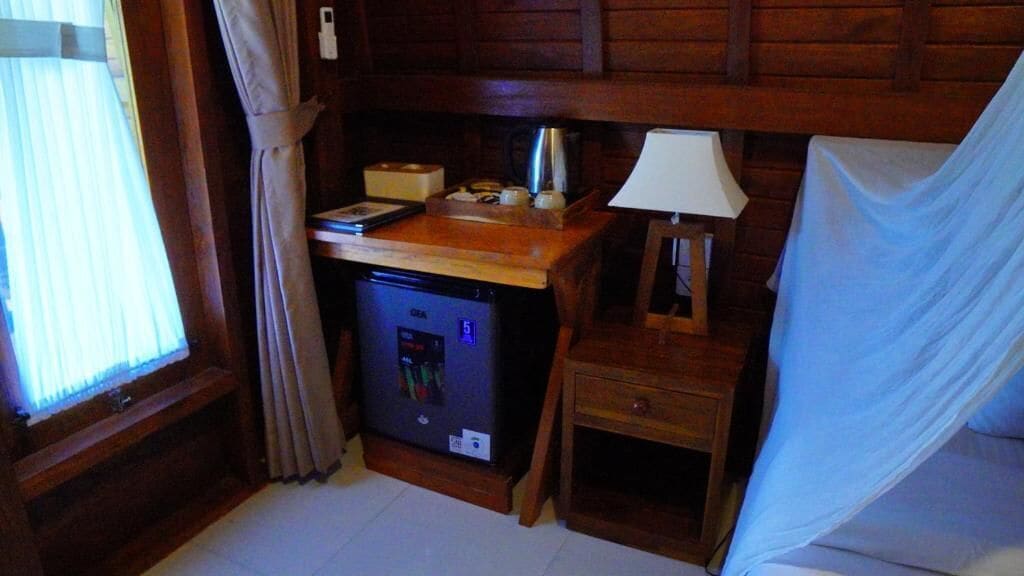 Habitación compacta con escritorio de madera, un refrigerador pequeño, una lámpara y una cama con dosel blanco.