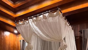 Een hemelbed met een witte spreideken en kussens, een netjes opgevouwen handdoek op het bed en houten lambrisering aan de wanden.