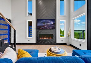 A modern living room with a fireplace, a large TV, and a view of the outdoors. - Mountain Views | EV Charger | Pool | Speakeasy (Broken Bow)