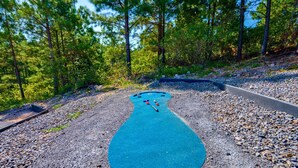 A blue shuffleboard court surrounded by a gravel path and dense forest. - Mountain Views | EV Charger | Pool | Speakeasy (Broken Bow)