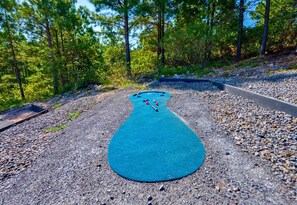 A blue shuffleboard court surrounded by a gravel path and dense forest. - Mountain Views | EV Charger | Pool | Speakeasy (Broken Bow)