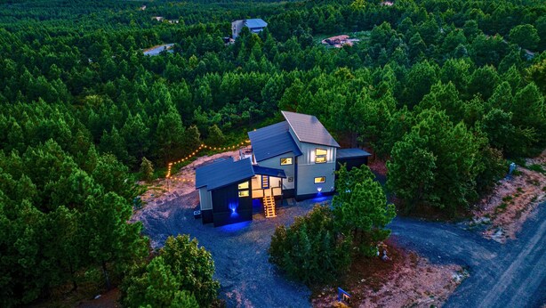 A modern house surrounded by dense forest, with a visible driveway and a small outdoor area with lights. - Mountain Views | EV Charger | Pool | Speakeasy (Broken Bow)