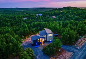 Moderni talo, jonka ympärillä on tiheä metsä, ja jolla on selkeästi näkyvissä oleva ajotie sekä pieni ulkoalue, jossa on valaistus.