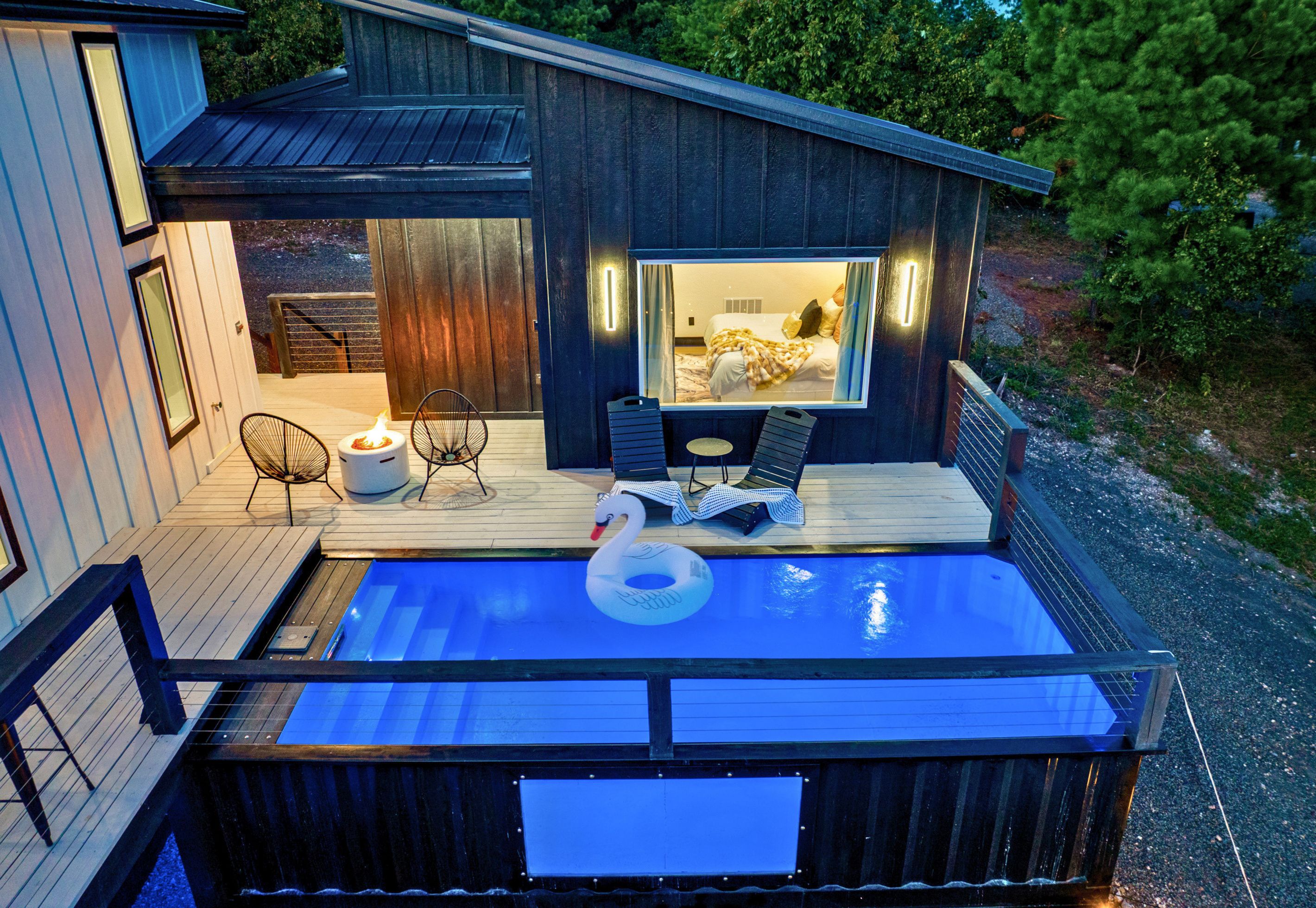 A hot tub with a swan float, a deck with chairs, and a view of a bedroom with a bed and curtains.