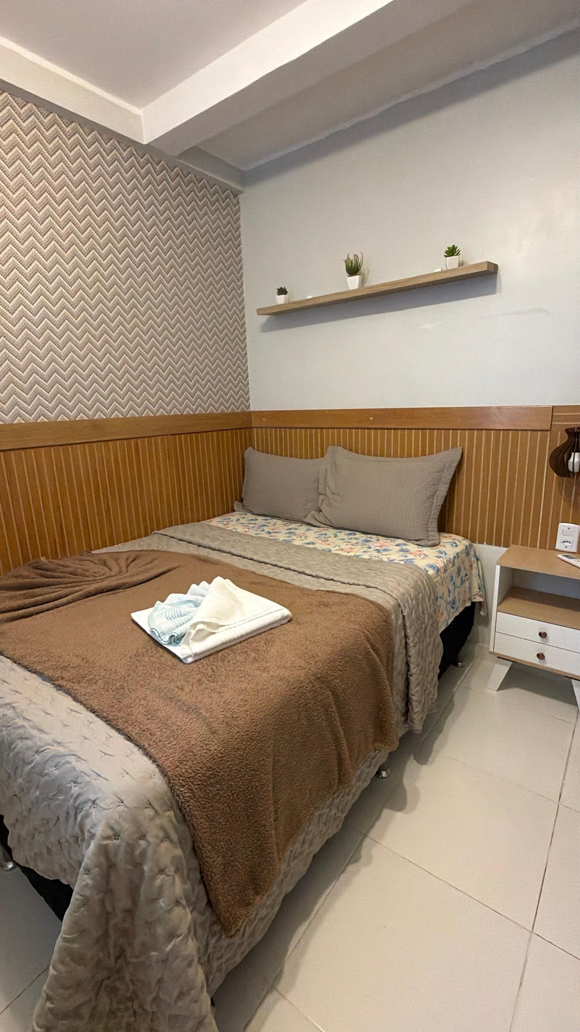 Un dormitorio ordenado con una cama, almohadas, una mesita de noche y una estantería con plantas.