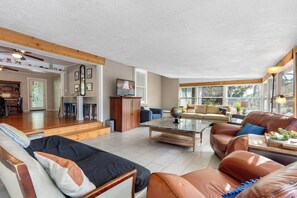 A spacious living area with a sofa, coffee table, and a television. There is a kitchen area with a dining table and chairs. The room has large windows allowing natural light. - Double Waterfront Estate; 12 Beds, Kayaks, Firepit (Tampa)