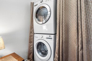 Una lavadora y secadora apiladas en una habitación con una cortina estampada y una lámpara sobre una mesita.