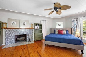 A bedroom with a fireplace, a bed with a blue bedspread, a green dresser, and a ceiling fan.