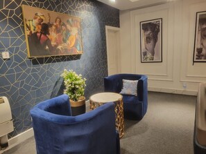 Ein Zimmer mit zwei blauen Sesseln, einem kleinen runden Tisch, einer Topfpflanze und einer Wand mit geometrischem Muster und Kunstwerken.