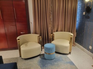 Ein Zimmer mit zwei Sesseln, einem kleinen Hocker, einem Kleiderschrank und einem Vorhang.