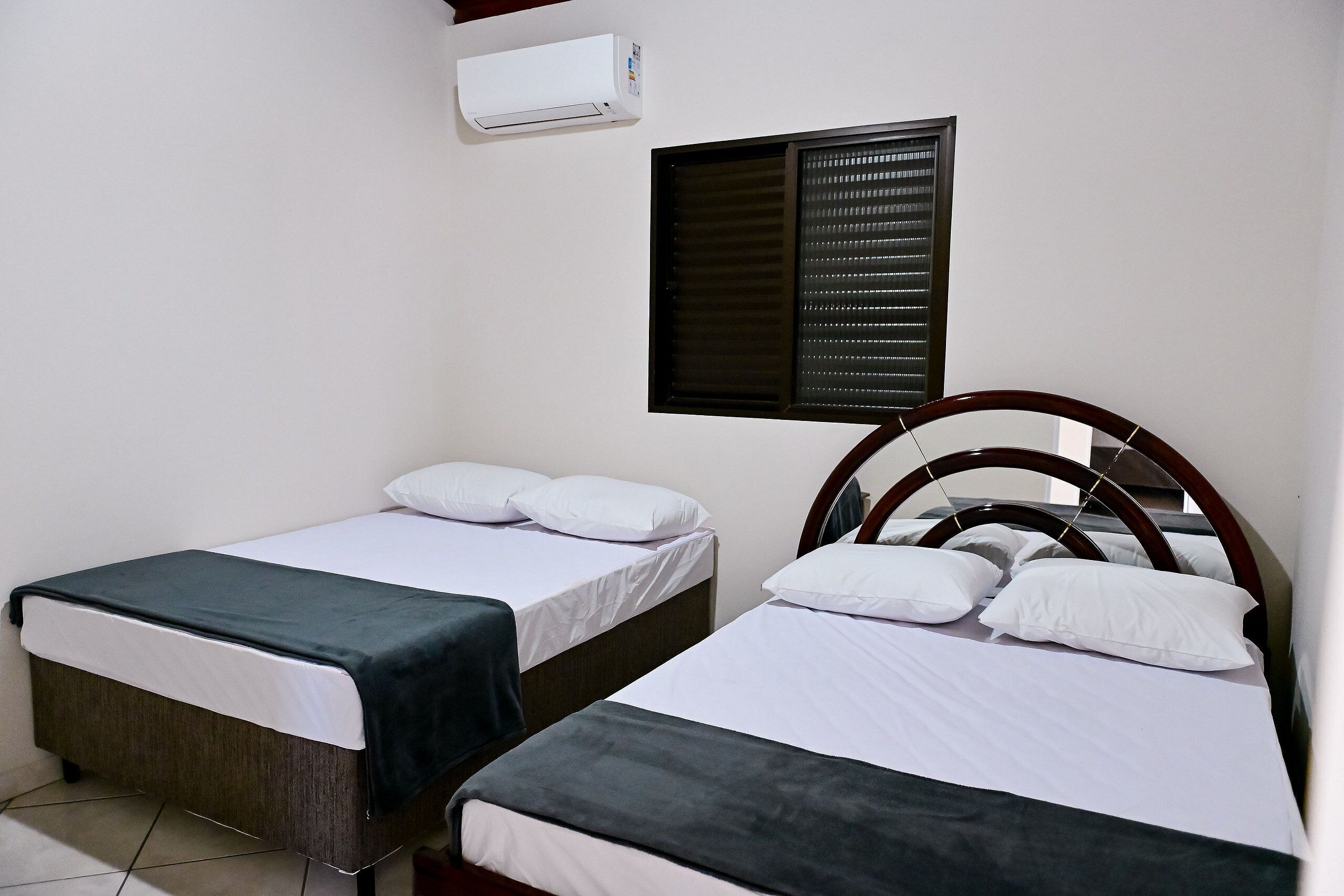 Une chambre d’hôtel avec deux lits simples, une fenêtre avec des stores, un appareil de climatisation et une tête de lit en bois.