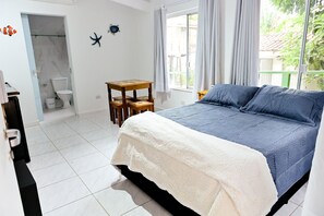 Ein Schlafzimmer mit einem Bett, einem kleinen Holztisch, einem weißen Stuhl, einer weißen Toilette und einem Fenster mit Vorhängen.