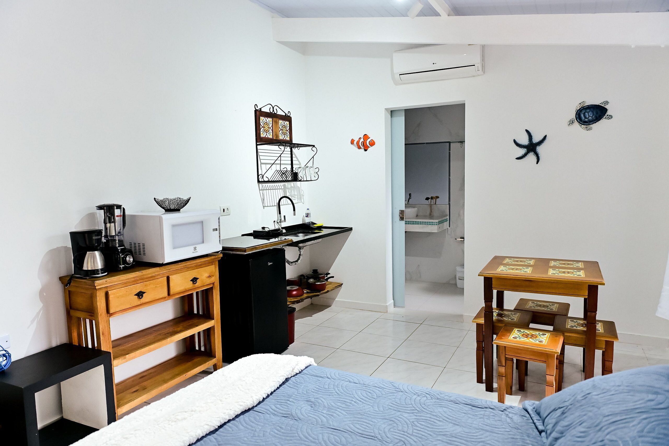 Quarto com cama, cómoda de madeira, kitchenette com micro-ondas e pequena mesa com cadeiras.
